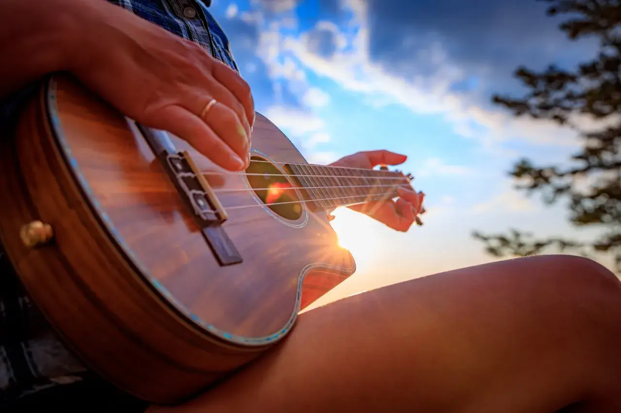 Dłonie grające na ukulele, słońce przebija się przez struny, tworząc magiczne akordy ukulele.