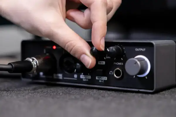 Dłoń kręci gałką miksera audio, regulując poziom sygnału. Widoczne złącza audio i wejścia mikrofonowe.